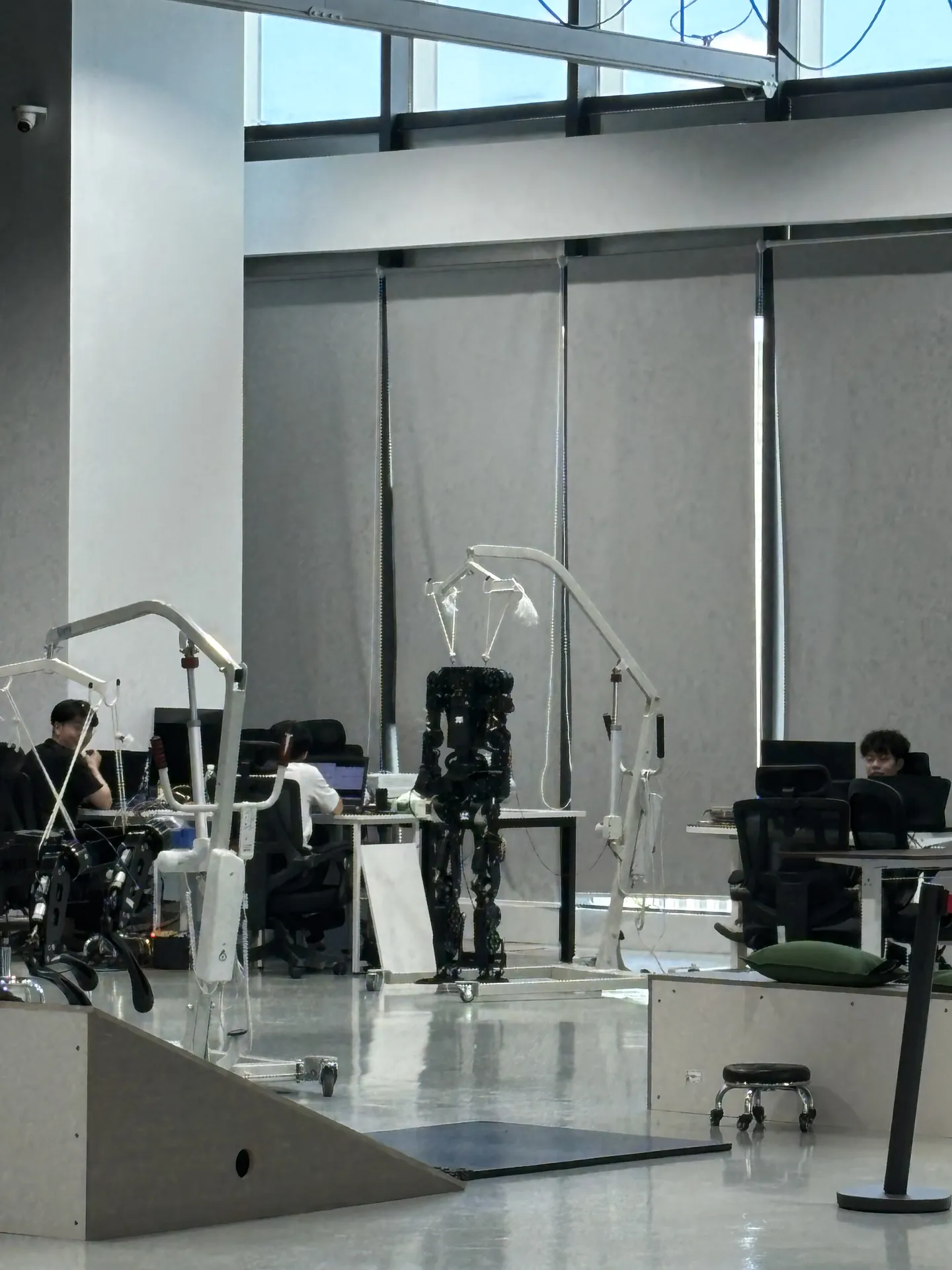 A black humanoid robot frame hanging in a lifting harness between desks